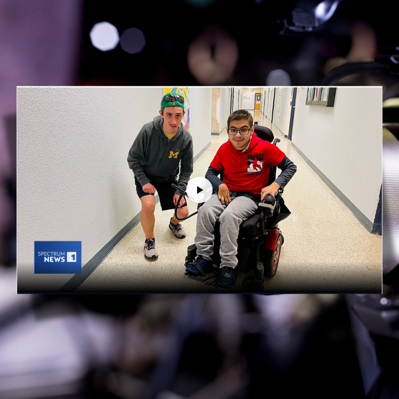   two students, one standing and one in a wheelchair, smiling at the camera. in the background is a 