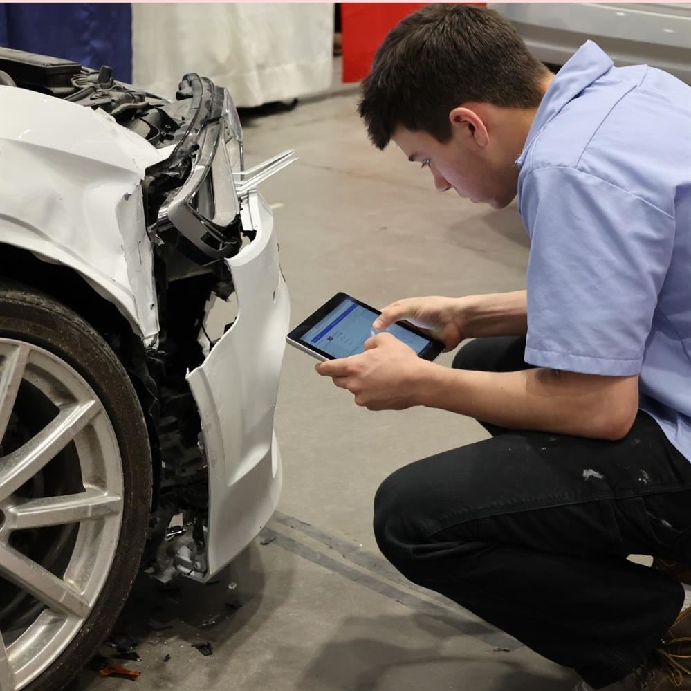  student kneeling by car tire, assessing damage to bumper